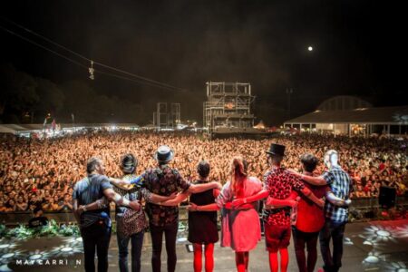 Un grupo de artistas en el escenario saludando a una gran multitud en un concierto al aire libre por.